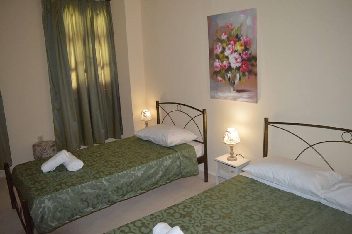 A hotel room with two single beds, each with a green bedspread and white pillows. There is a painting of flowers on the wall above the beds.