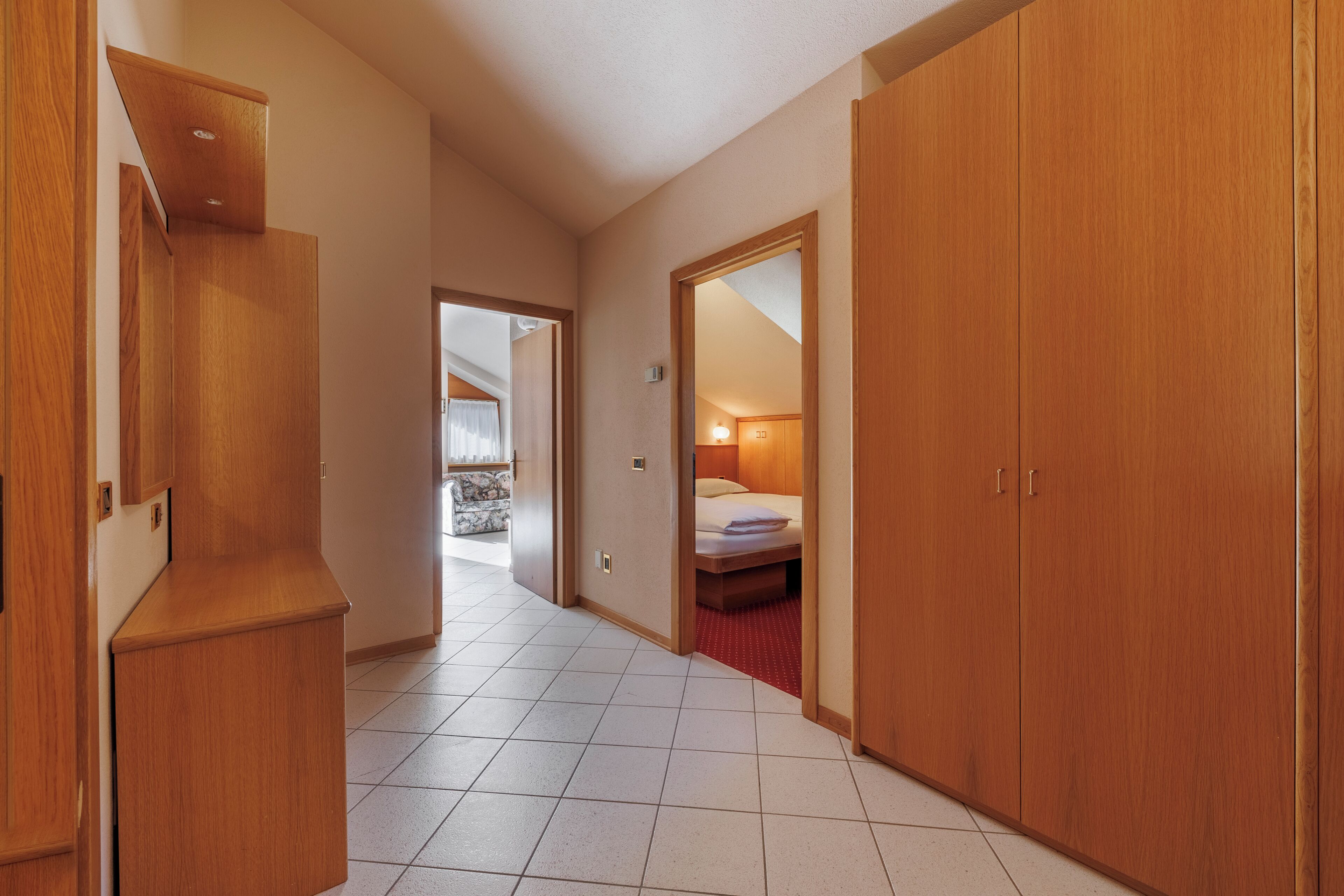 Corredor de hotel com armários de madeira, uma cama e um banheiro visível por uma porta aberta.