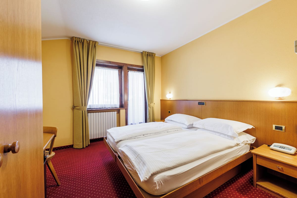 Habitación de hotel con dos camas, cabecera de madera, mesita de noche, una silla, ventana con cortinas y un radiador.