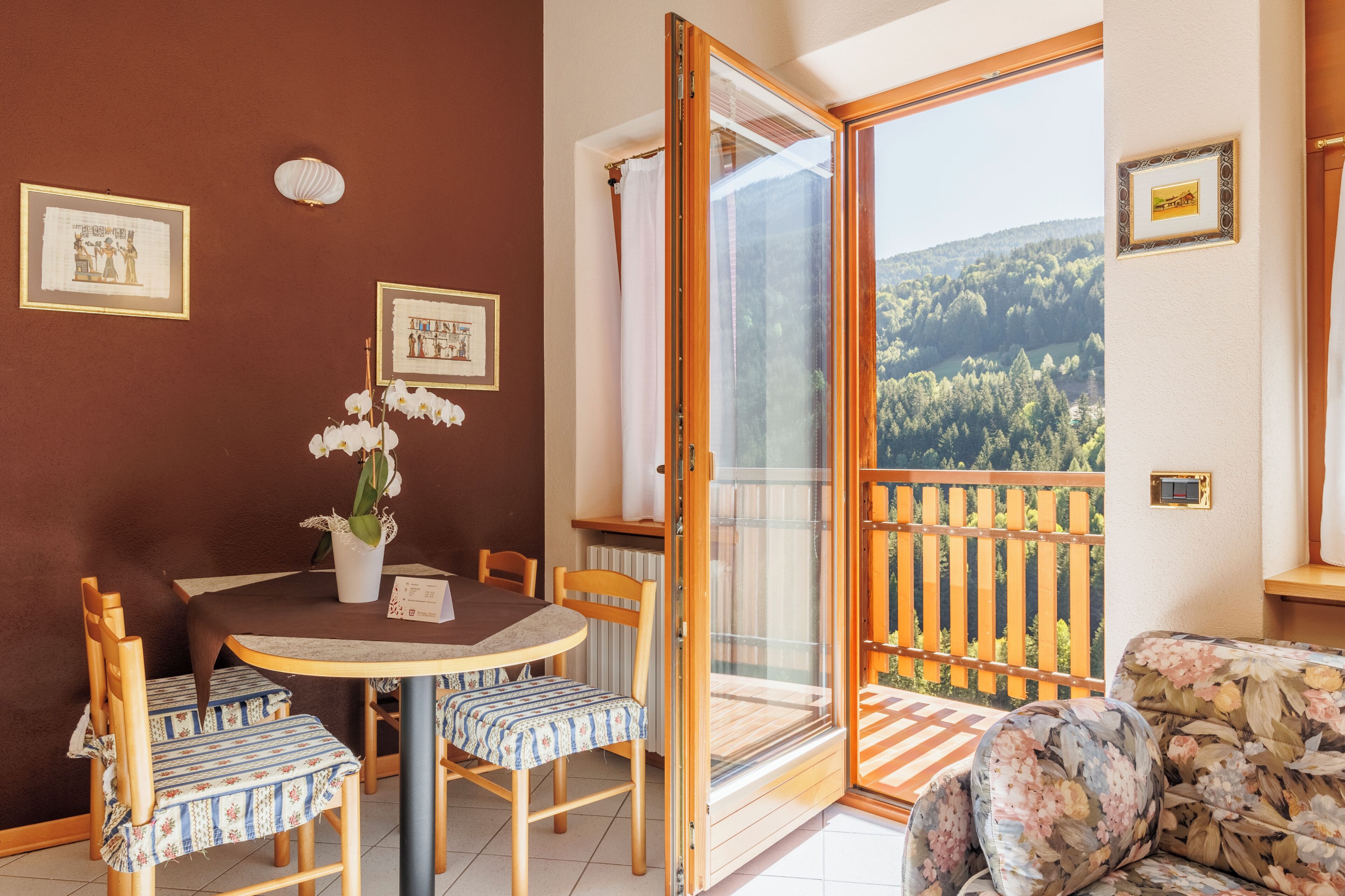 Et værelse med et spisebord, stole, en stueplante, en sofa og en balkon med udsigt over et skovområde.