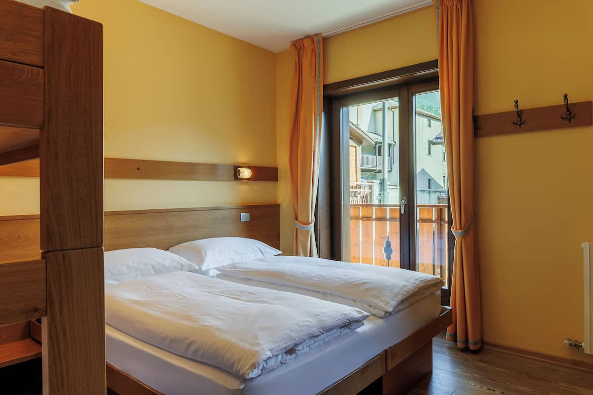 Cama doble con cabecera y piecera de madera, mesita de noche, ventana con cortinas y vistas a un balcón.