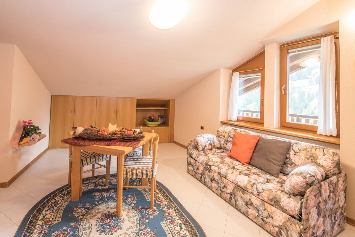 Un salón acogedor con una mesa redonda, sofá floral y vistas al exterior a través de una ventana.