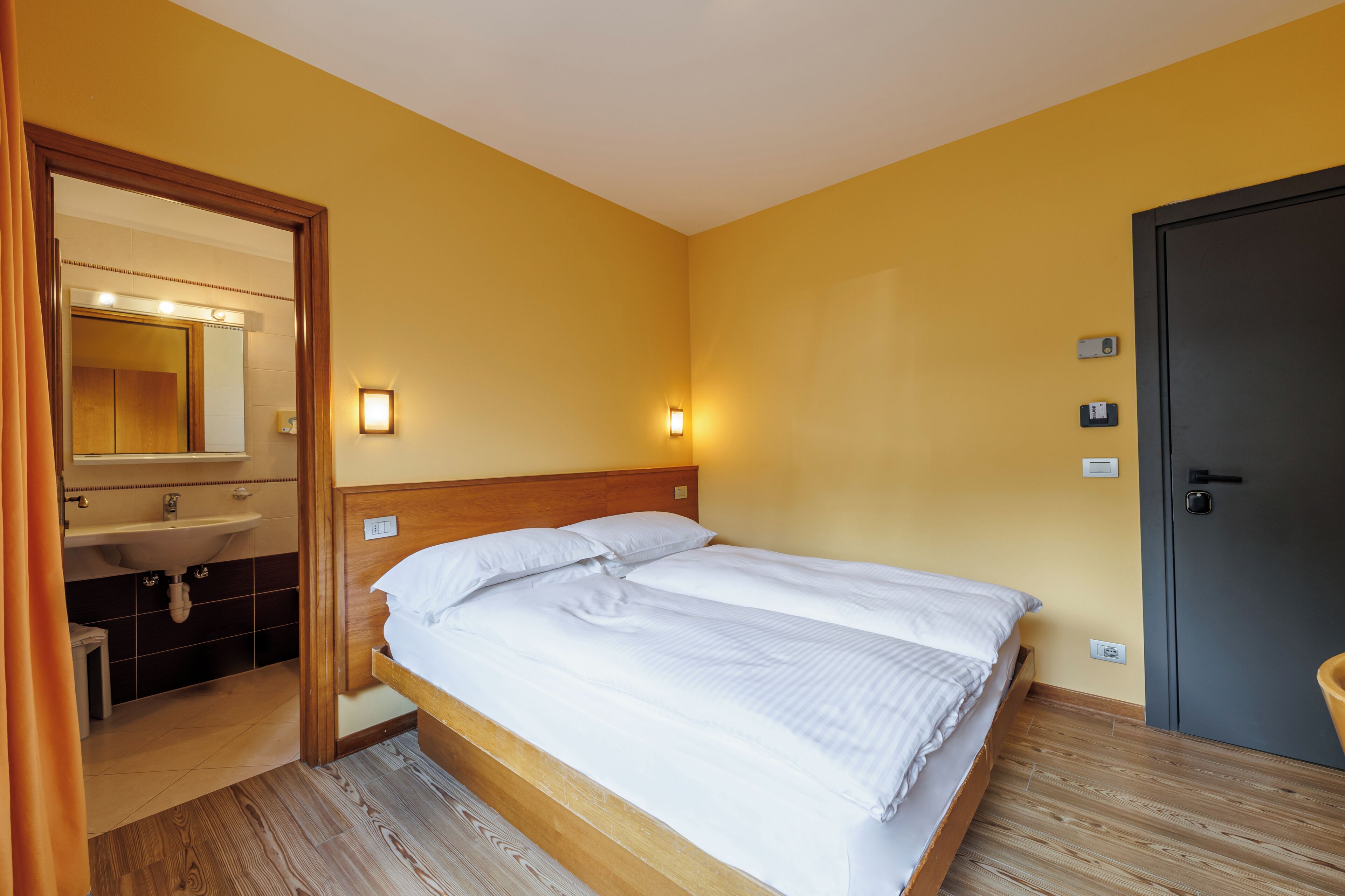 Habitación de hotel con una cama de madera, un baño con lavamanos y espejo, y una puerta con cerradura.