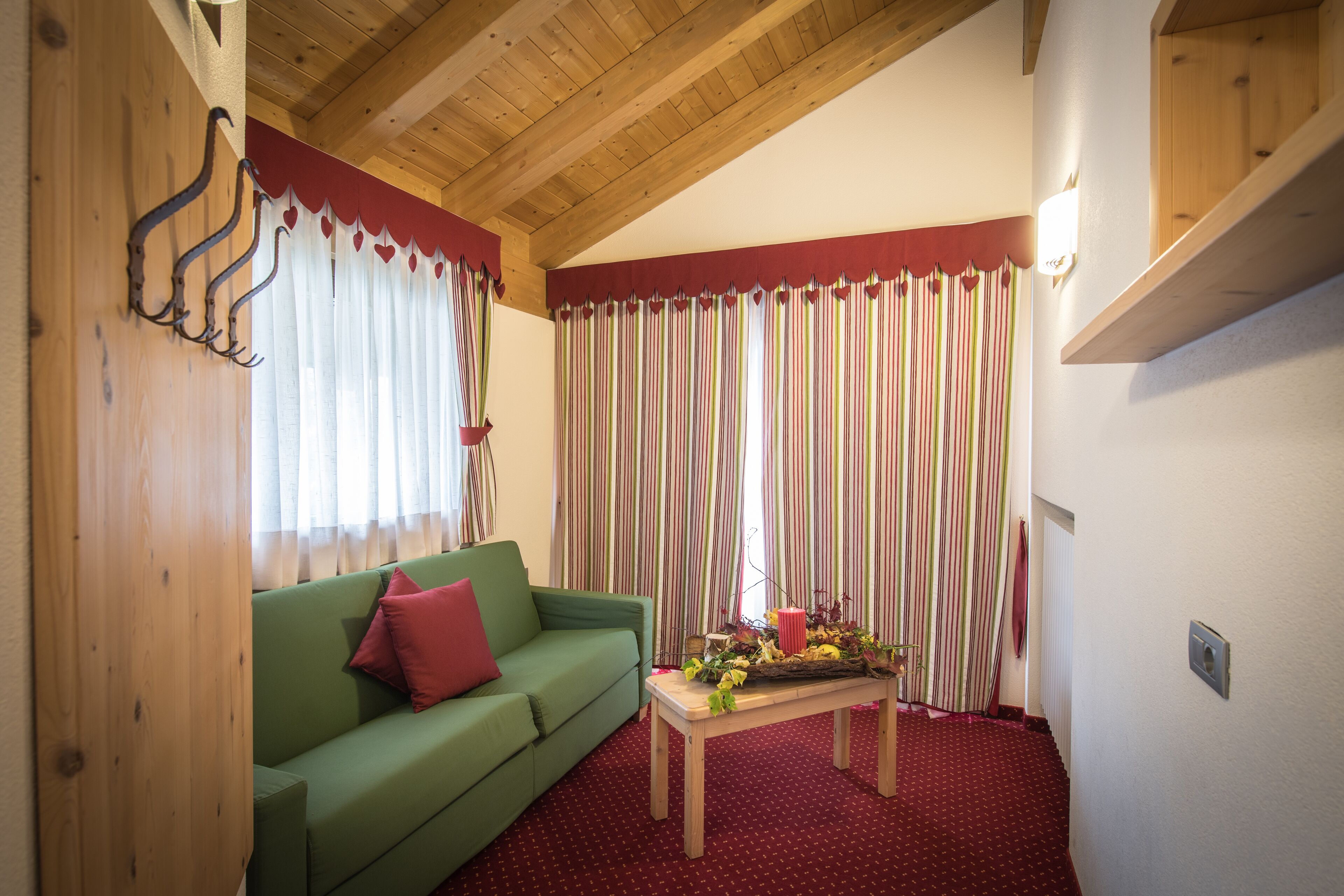 Una habitación con techo de madera, un sofá verde con un cojín rojo, una mesa con un arreglo floral y cortinas a rayas.