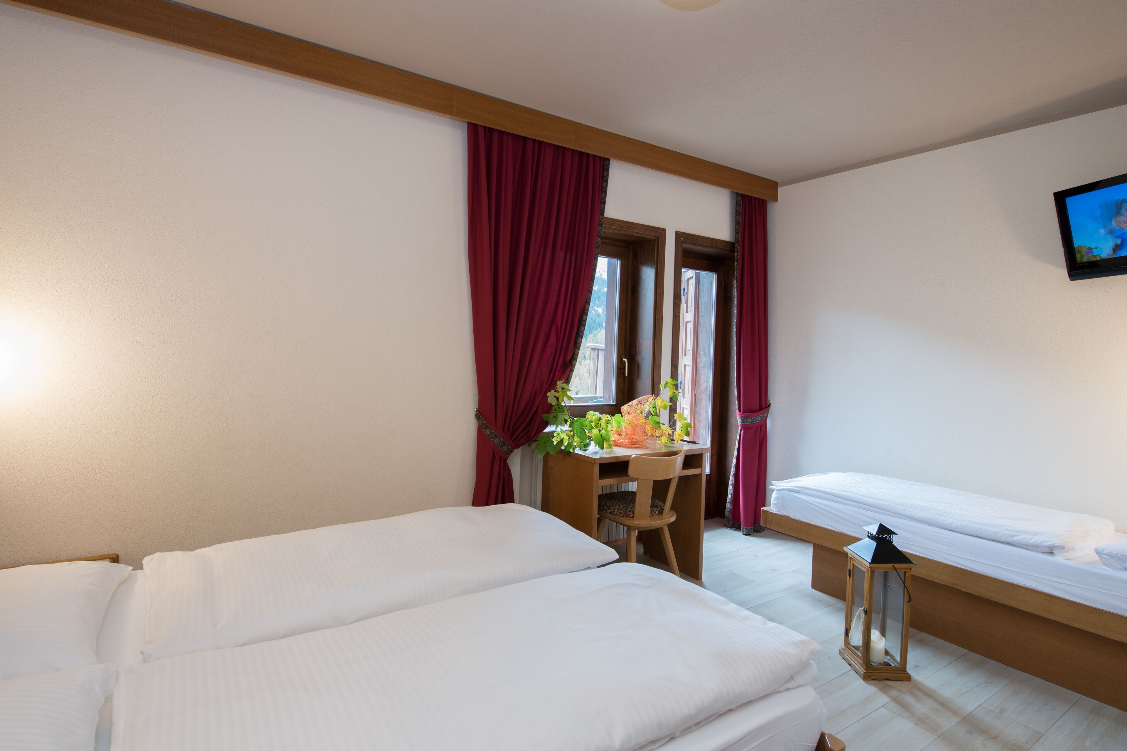 Habitación de hotel con dos camas, un escritorio y una silla, una ventana con cortinas rojas y un televisor en la pared.