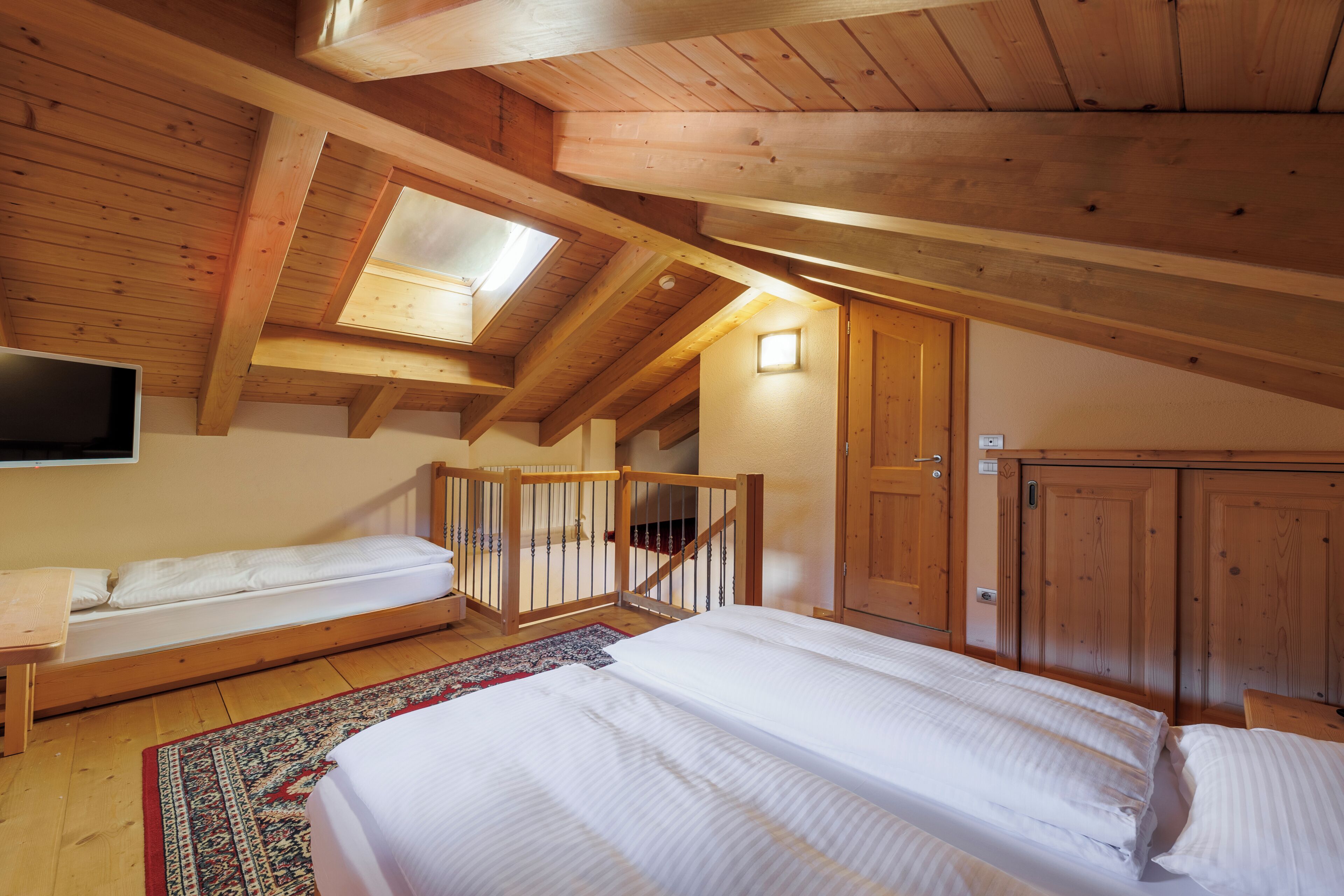 A room with a loft bed, a lower bunk bed, wooden beams, a skylight, a staircase, and a wardrobe.