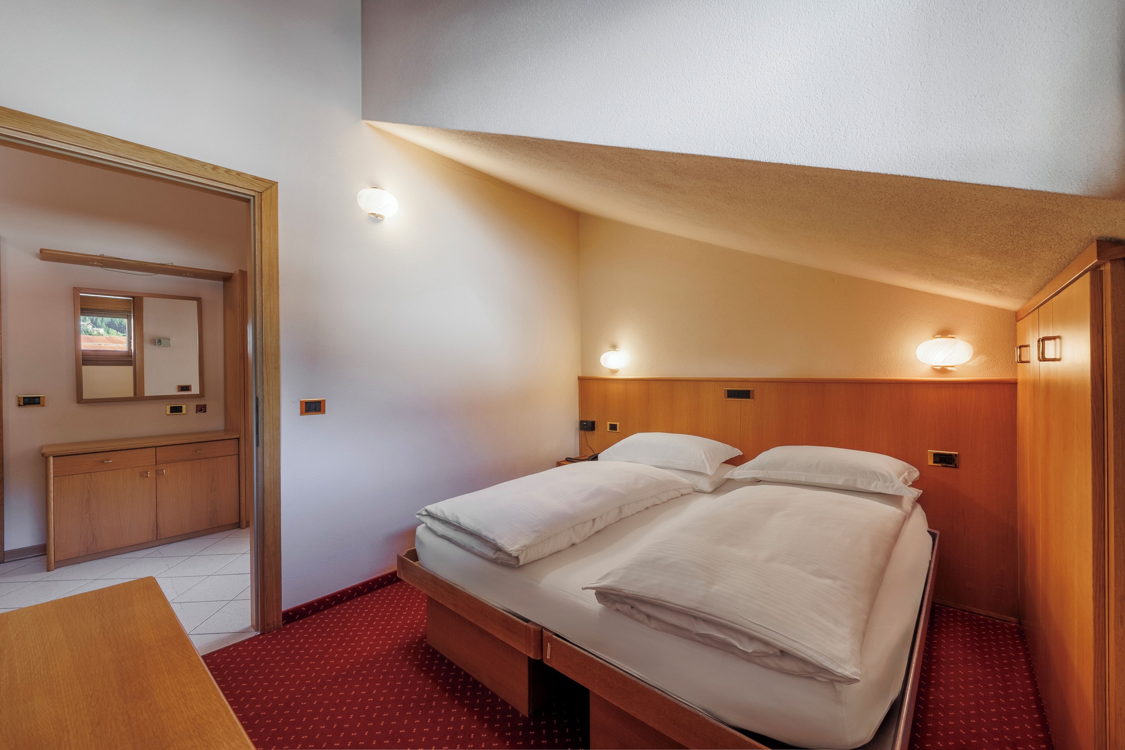 Et lille hotelværelse med en enkeltseng, et træskab, et skrivebord og et spejl på væggen.