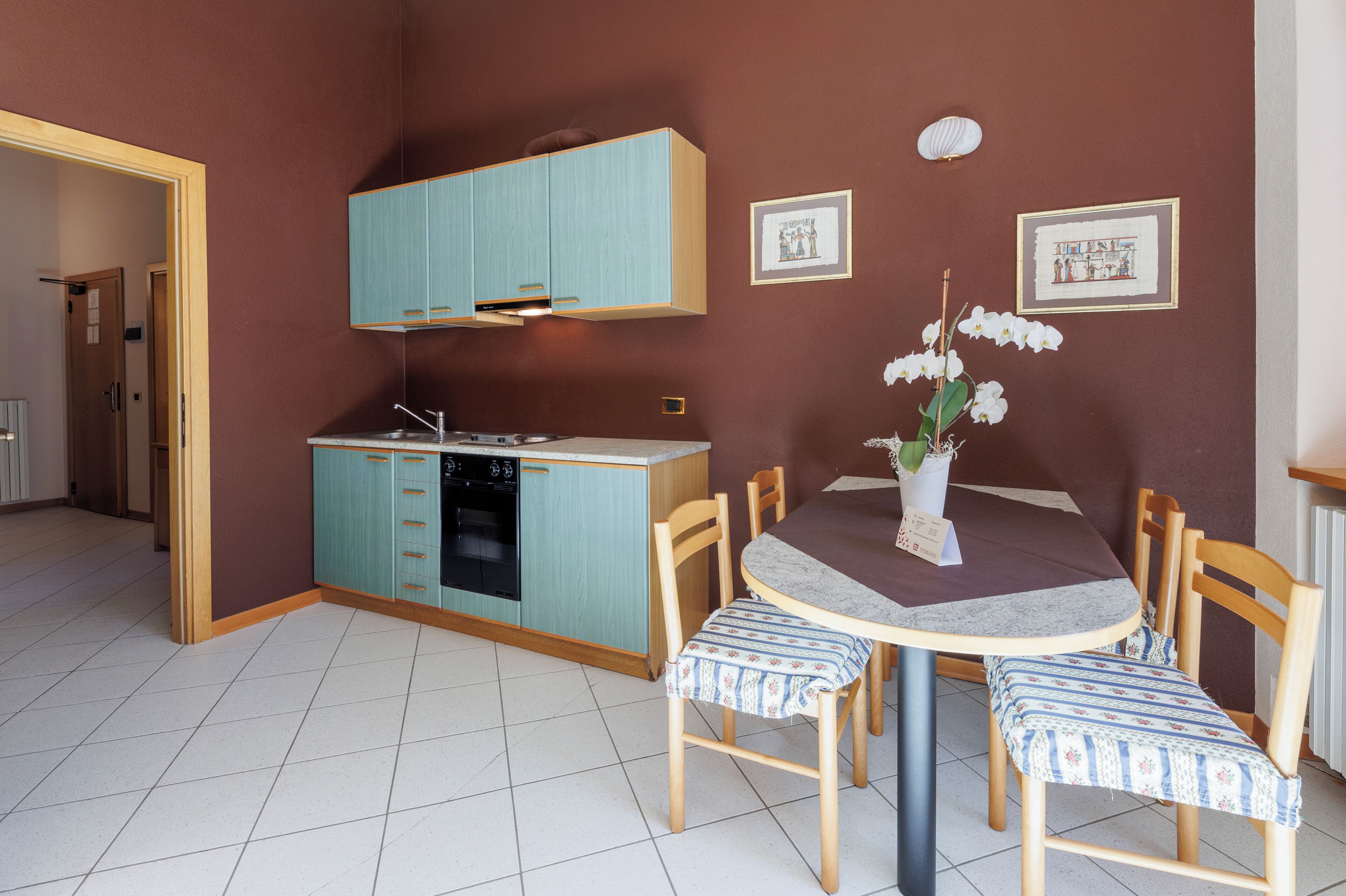 Een compacte keuken met blauwgroene kastjes, een eettafel met stoelen, en een vaas met bloemen.