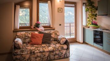 A living room with a floral sofa, a kitchen with a window, and a balcony with a view of trees.
