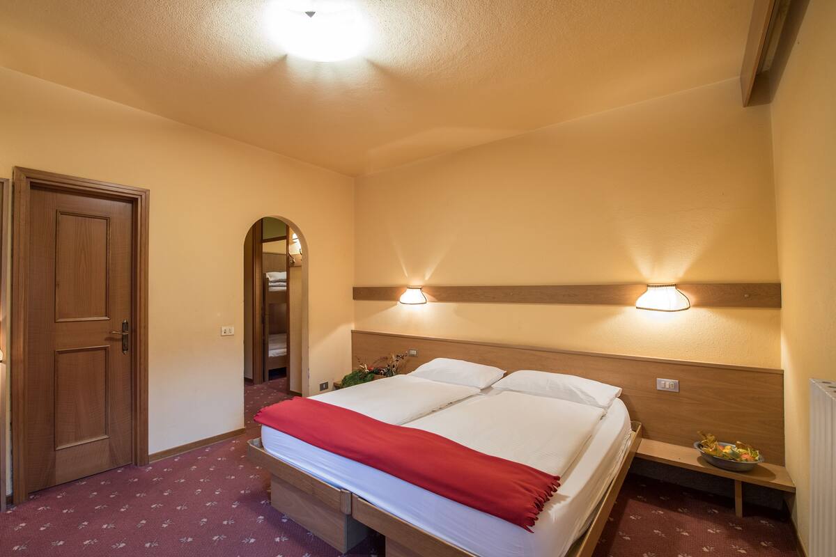 Habitación de hotel con cama, cabecera de madera, dos luces fijadas a la pared, una puerta y una mesita de noche con un cuenco.