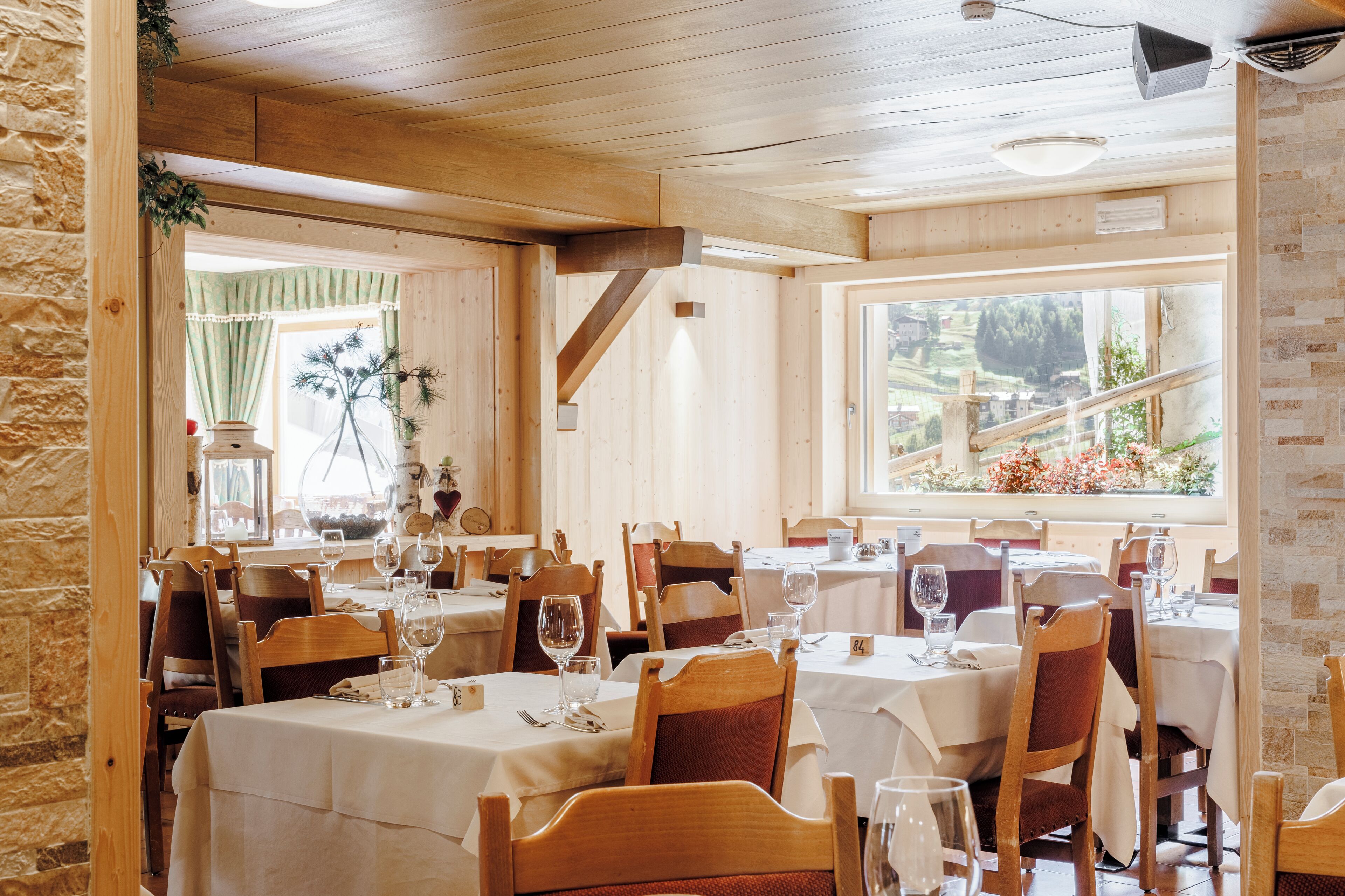 A restaurant with wooden interiors, multiple tables, and chairs, set against a stone wall and a window with a view of a garden.