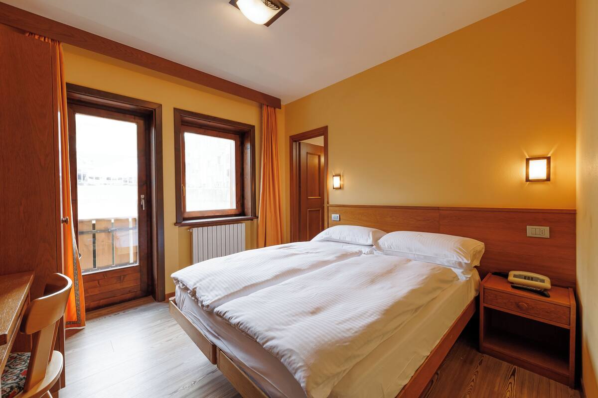 Un dormitorio con dos camas, cabeceros de madera, una mesita de noche con lámpara y una ventana con cortinas.