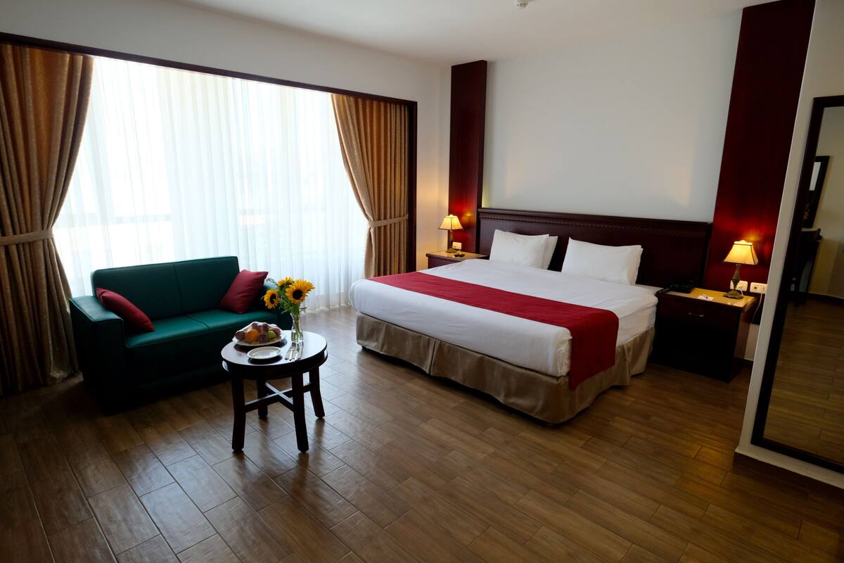 A hotel room with a bed, a green sofa, a small table with a vase of flowers, and a nightstand with a lamp.