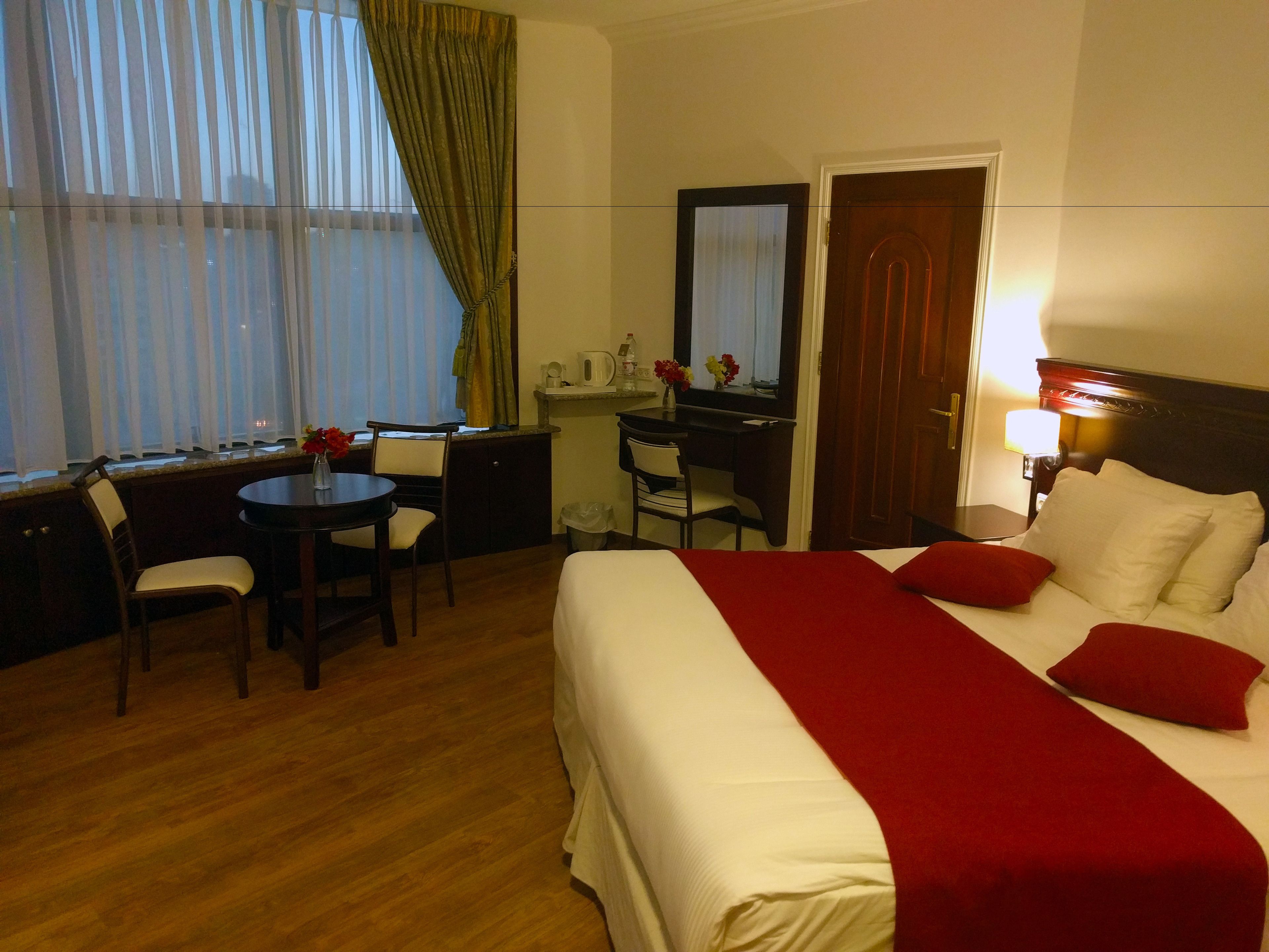 A hotel room with a bed, a desk, a chair, a small table, a mirror, a lamp, and a window with curtains.