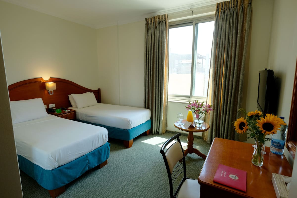 A hotel room with two beds, a desk with a chair, a table with a vase of flowers, and a window with curtains.