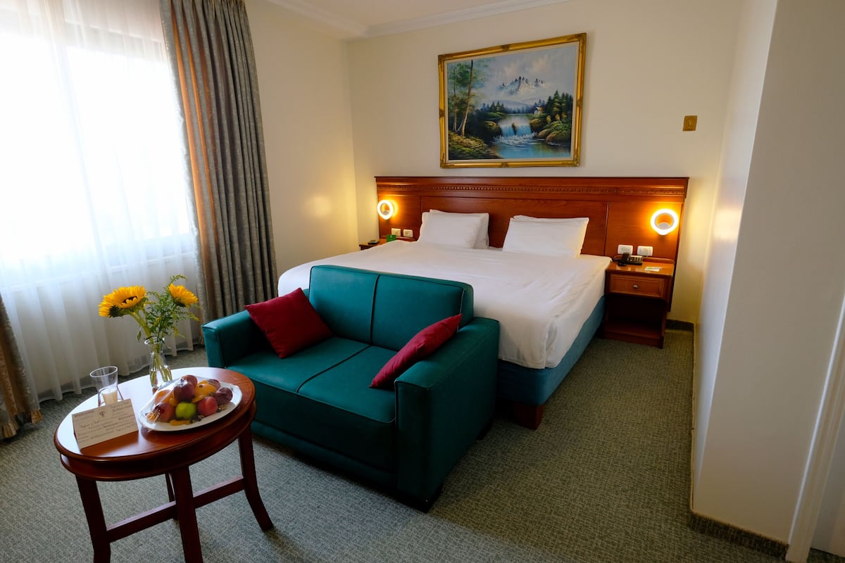 A hotel room with a bed, a green sofa, a small table with a fruit plate and a vase of sunflowers, and a painting above the bed.