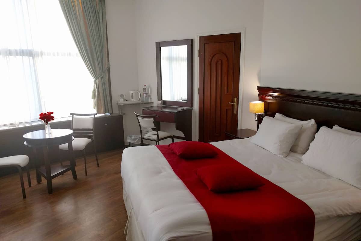 A hotel room with a bed, a desk, a chair, a small table with a vase of flowers, and a window with curtains.