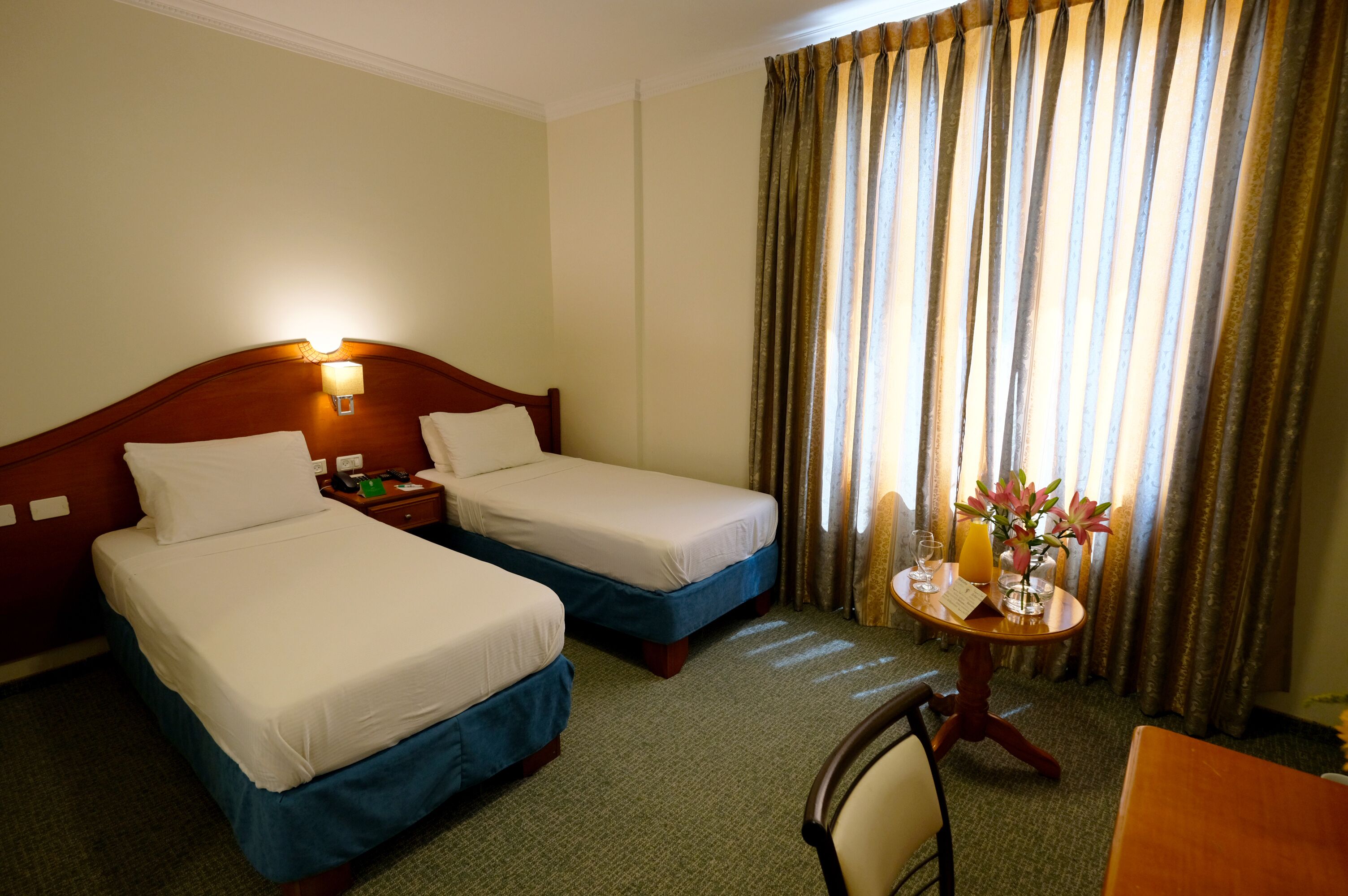 A hotel room with two single beds, a bedside table with a lamp, a small table with a vase of flowers and a glass of orange juice, and a chair.
