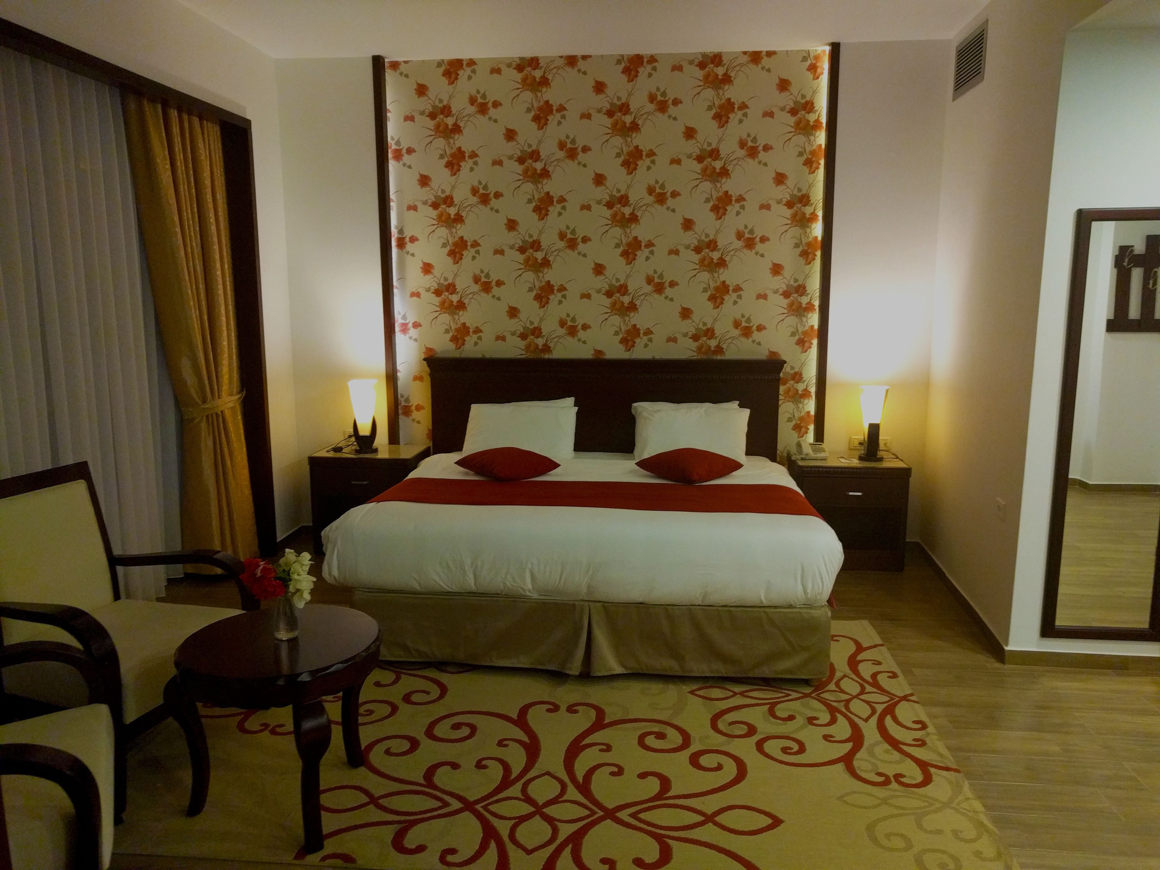 Una habitación de hotel con una cama grande, dos mesitas de noche con lámparas, una pequeña mesa redonda con un jarrón y una silla.