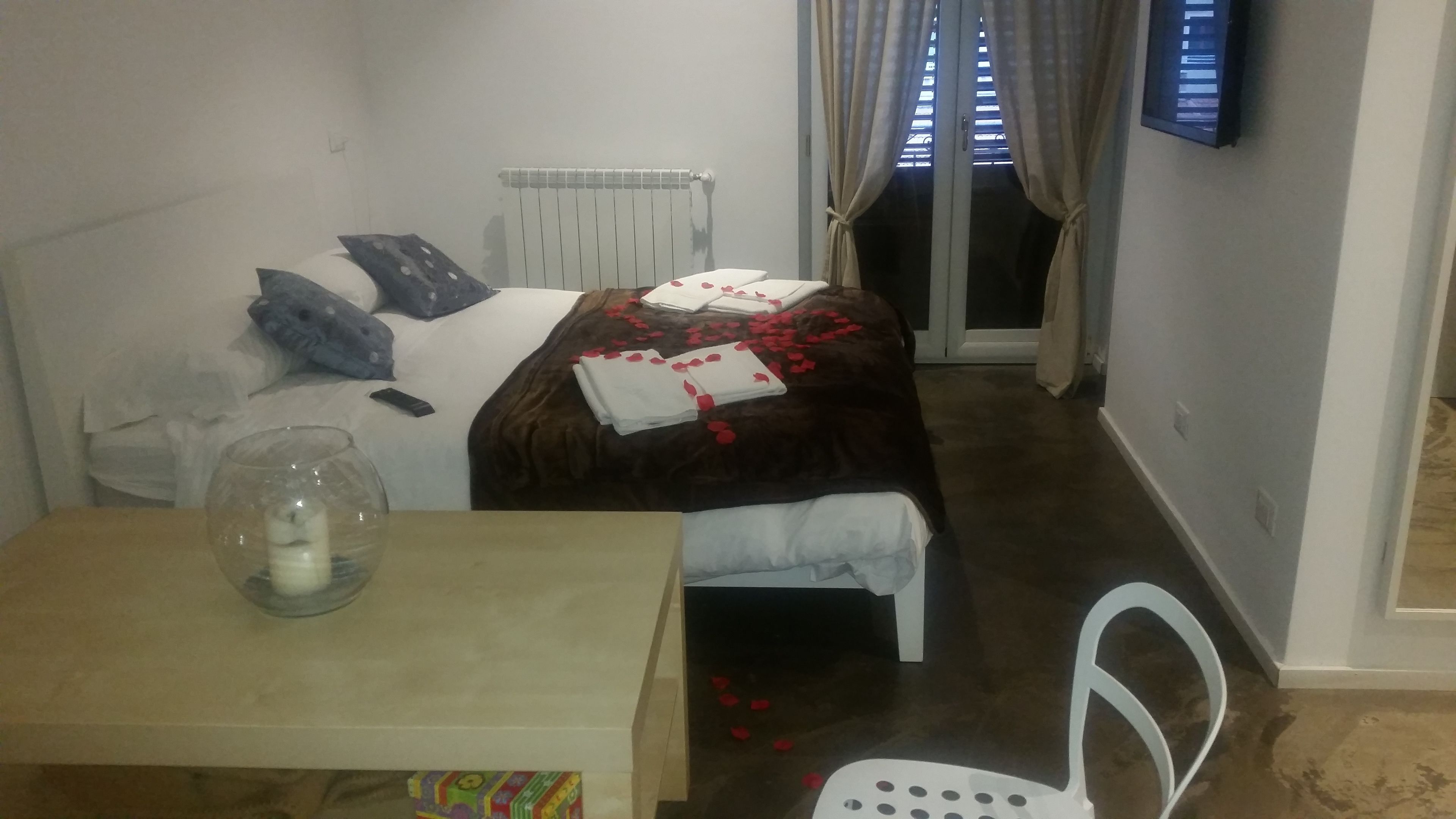 A small, neatly arranged bedroom with a bed, a bedside table with a glass, a chair, and a TV on the wall.