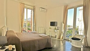 A hotel room with a bed, a desk, a chair, a TV, and large windows offering a city view. - Palermo Rooms (Palermo)
