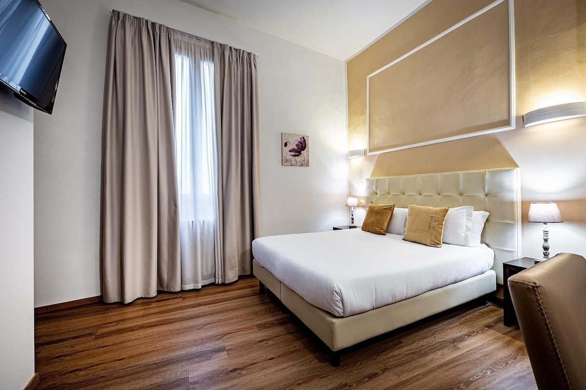 Ein Hotelzimmer mit einem großen Bett, einem flachbildfernseher an der Wand, einem Fenster mit Vorhängen, einem Nachttisch mit Lampe und einer Wandleuchte.