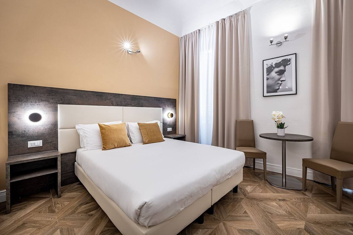 Ein Hotelzimmer mit einem großen Bett, Nachttischen, einem runden Tisch mit Blumen in einer Vase und einem gerahmten Bild an der Wand.