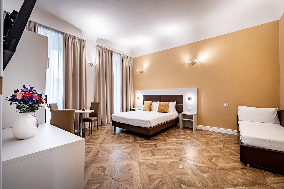 Ein Hotelzimmer mit Bett, Nachttischen, Schreibtisch, Sessel und Blumen in einer Vase.