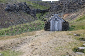 Un pequeño edificio de piedra con techo a dos aguas, situado en una zona montañosa con un camino de tierra que lo lleva a él.