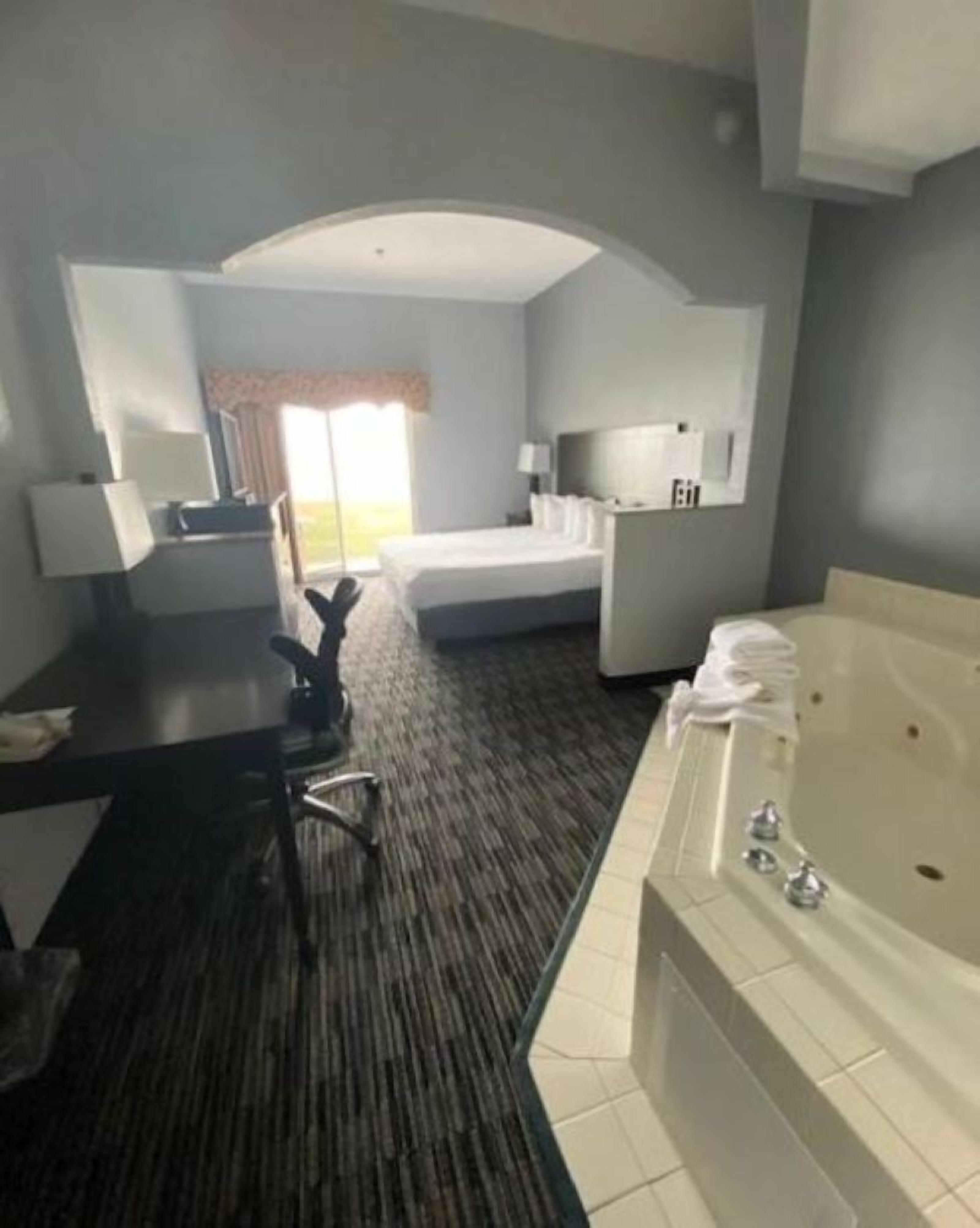 A hotel room with a bed, a desk, a chair, a desk lamp, a TV, a bathroom with a bathtub, and a window with curtains.