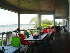 A covered outdoor dining area with a view of a lake, featuring a long table with chairs and potted plants. - Lakeside Resort and Conference Center (Houghton Lake)