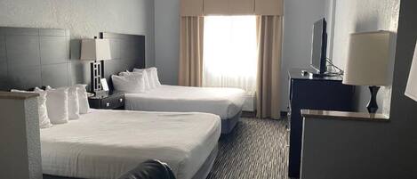 Ein Hotelzimmer mit zwei Betten, einem Schreibtisch mit Lampe, einem Fernseher und einem Fenster mit Vorhängen.