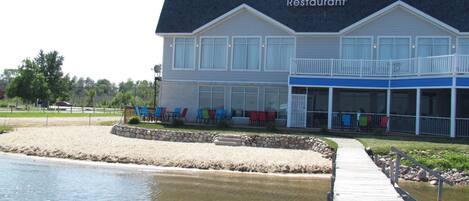 A restaurant with a covered outdoor seating area, a sandy beach, and a wooden dock leading to the water.