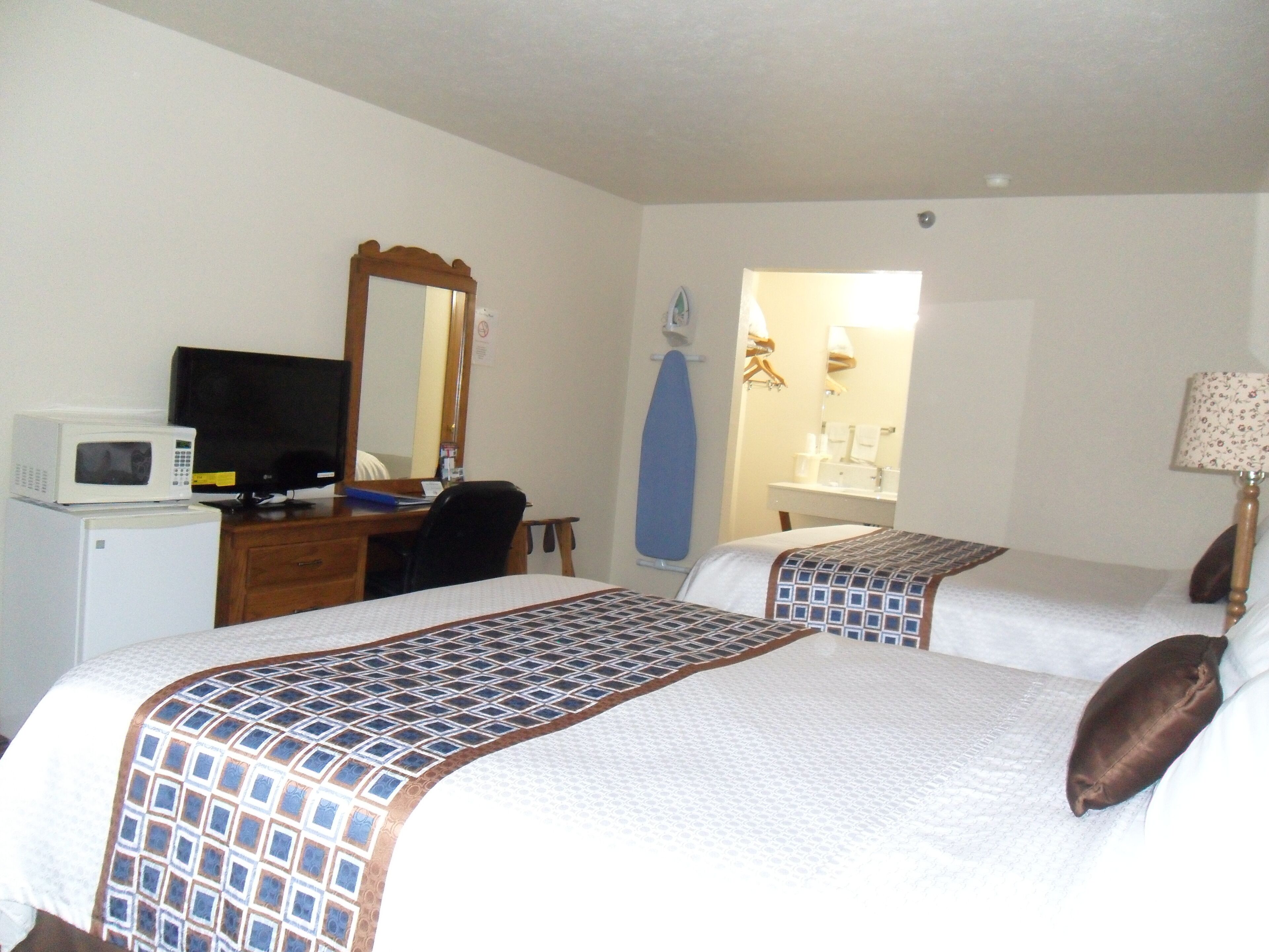 A hotel room with two beds, a desk with a computer and a microwave, a mirror, and a bathroom visible through an open door.