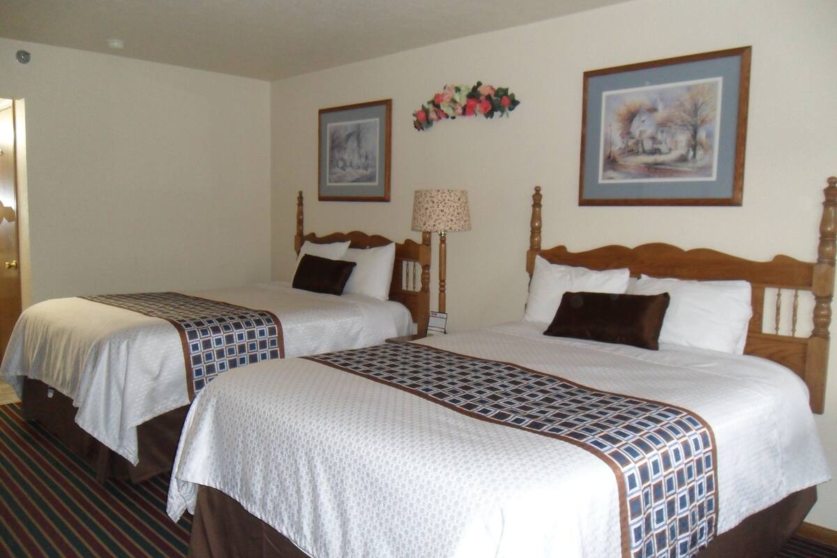 A hotel room with two beds, each with a wooden headboard, white bedding, and a patterned bedspread. There is a framed picture on the wall above the beds and a lamp on the nightstand.