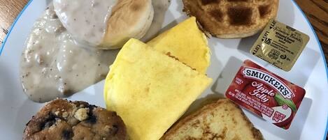 A plate with breakfast items including biscuits, waffles, toast, apple jelly, and a packet of whipped cream.
