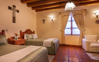 Habitación de hotel con dos camas, cabecera de madera, mesita de noche, lámpara, ventana con cortinas y un pequeño sofá.