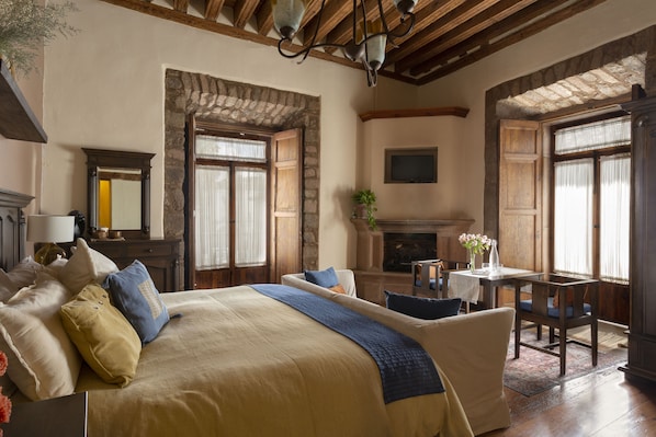 A bedroom with a large bed, wooden beams, a fireplace, and a dining area with a table and chairs. - Hotel Casa del Naranjo (Patzcuaro)