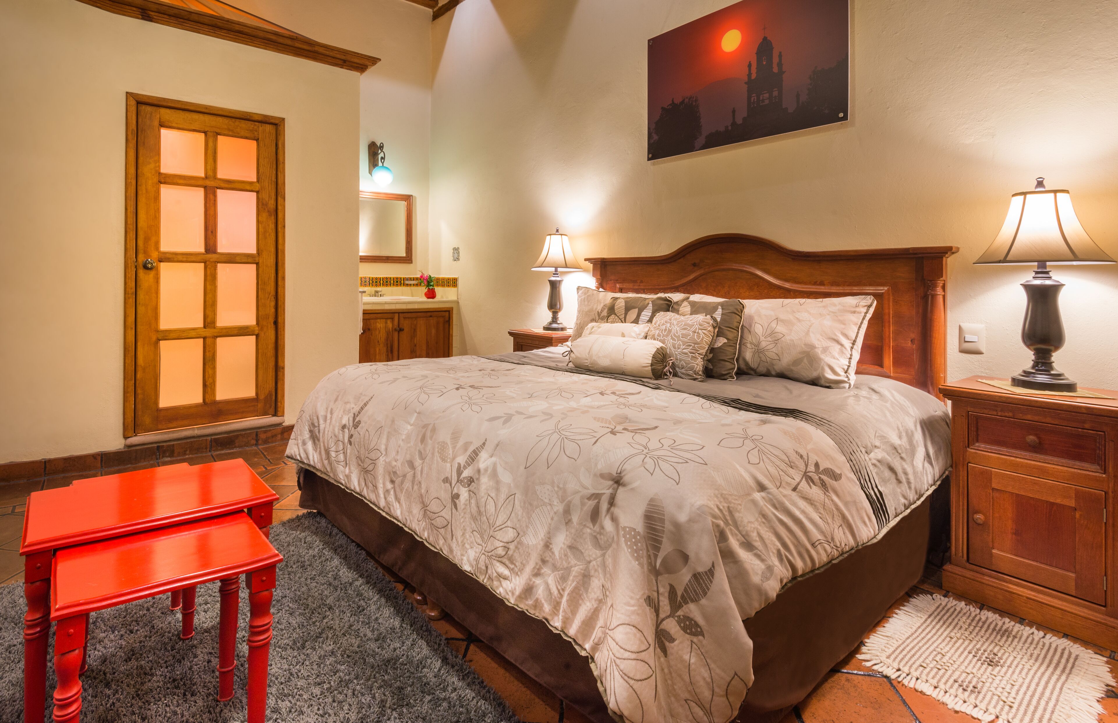 A bedroom with a large bed, two bedside lamps, a wooden nightstand, a red table, and a door with frosted glass panels.