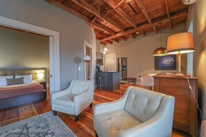 Una habitación con una cama, dos sillones de cuero blanco, techo de madera y una cocina al fondo.