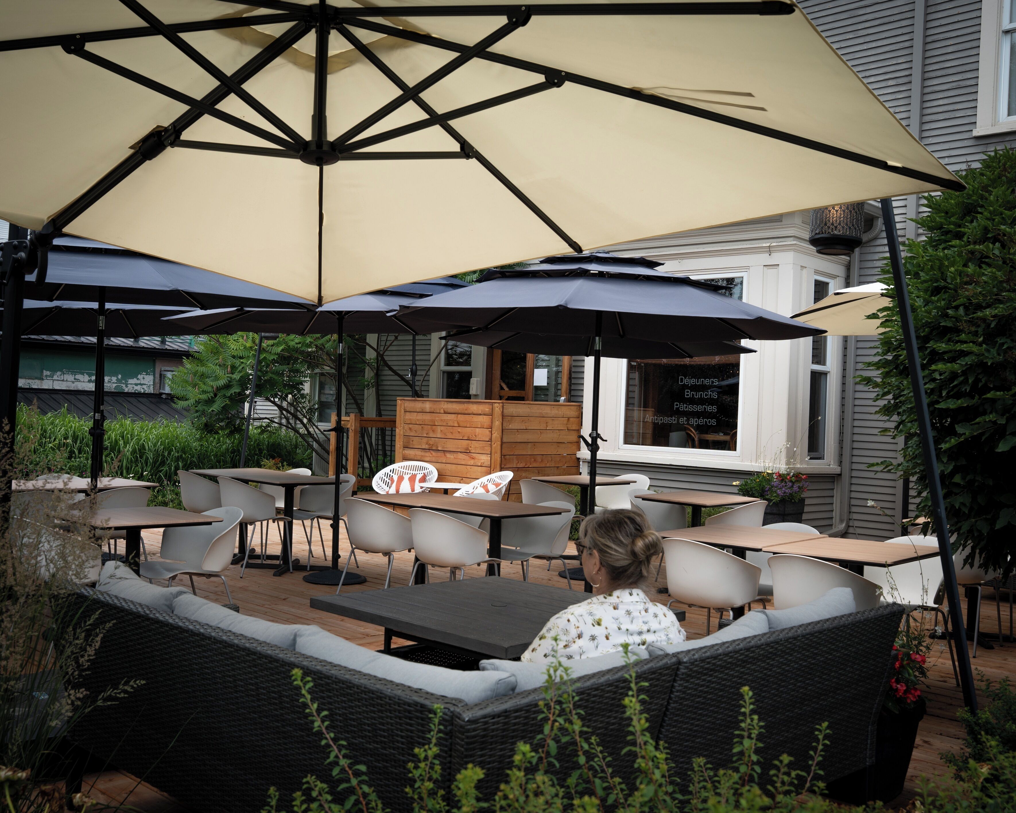 An outdoor seating area with tables and chairs under large umbrellas, a wooden deck, and a building with a window displaying a menu.