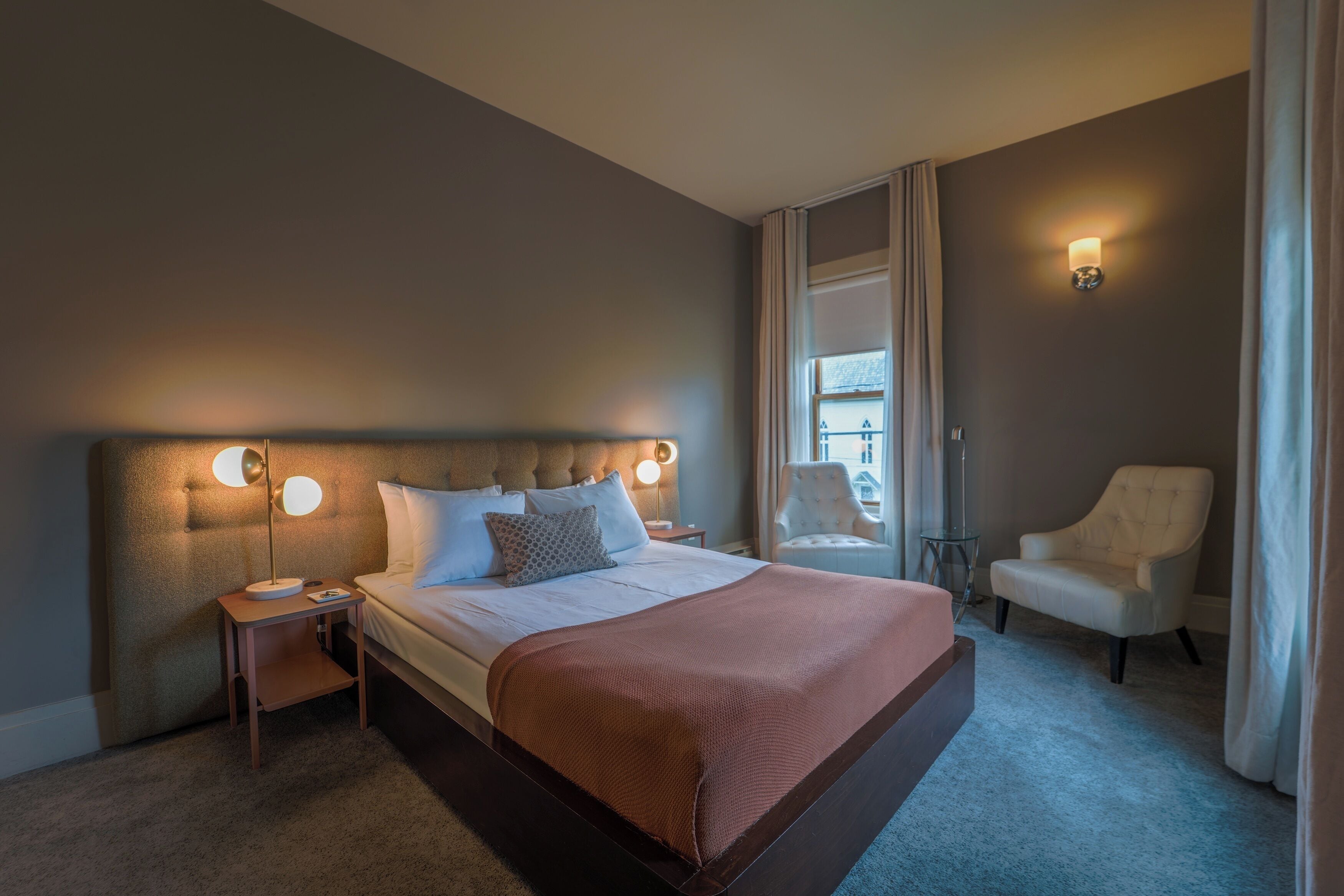 A hotel room with a large bed, a chair, a nightstand, and a window with curtains.