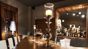 Una larga mesa de madera para comer con velas, copas y una zona de bar al fondo.