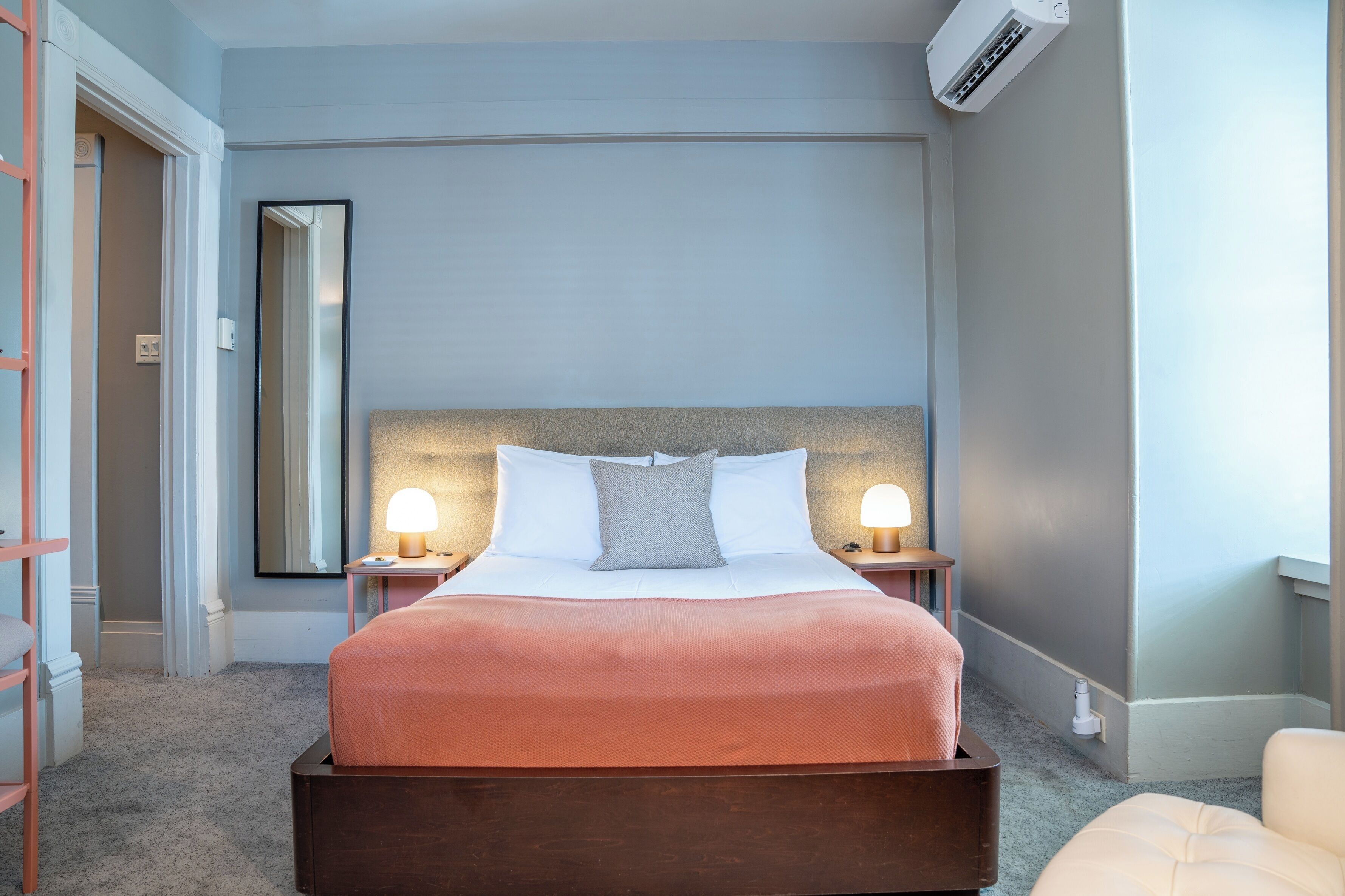 A hotel room with a bed, bedside lamps, a large mirror, and an air conditioning unit.