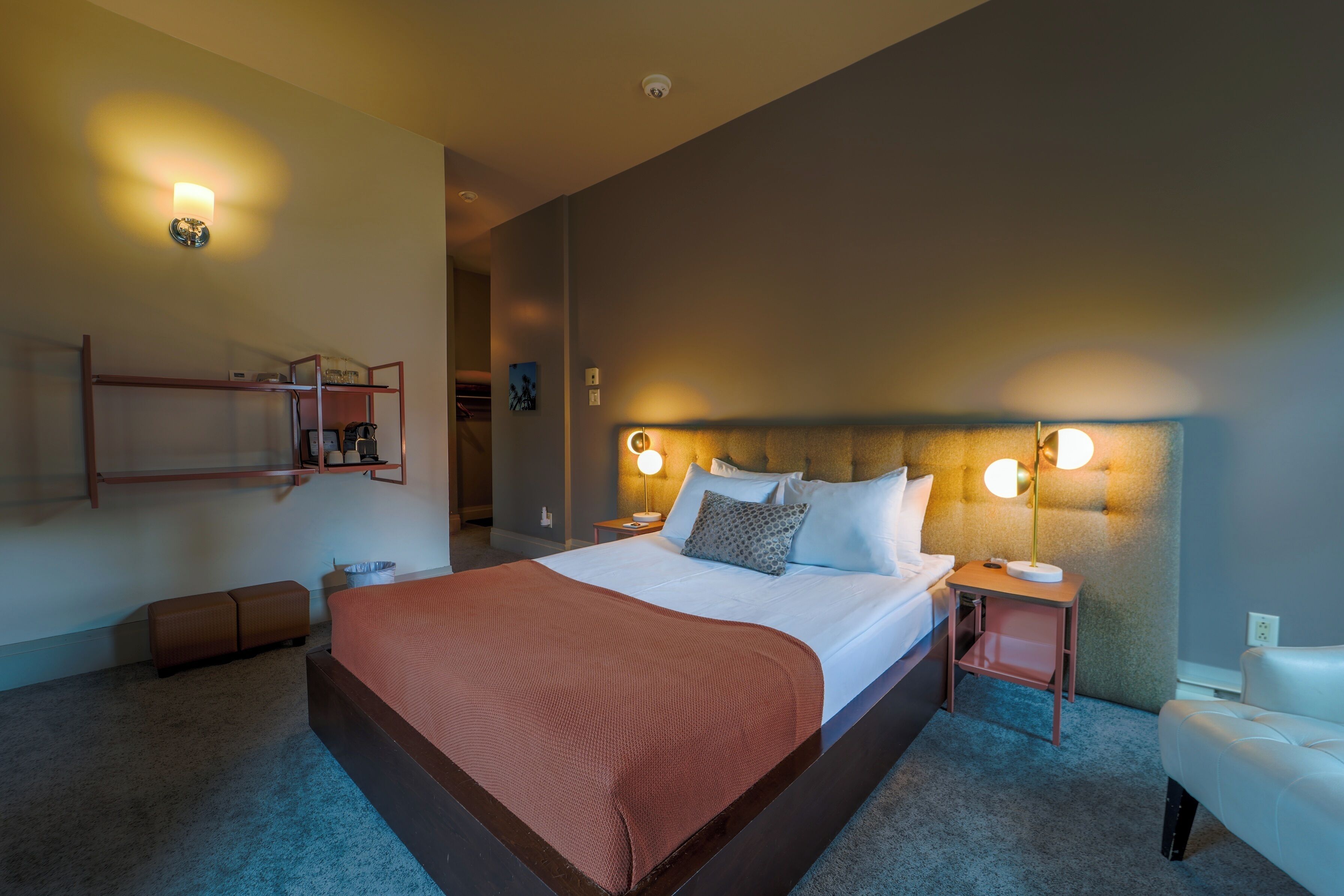 Ein Hotelzimmer mit einem großen Bett, Nachttischen, einem an der Wand befestigten Regal und einer Tür zu einem weiteren Raum.