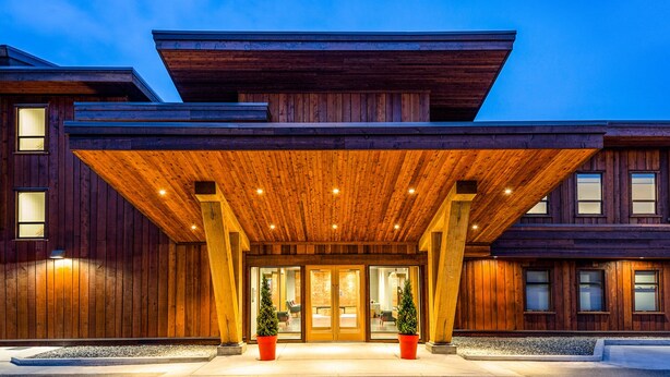 Un edificio moderno con fachada de madera, grandes puertas de vidrio en la entrada y un área de acceso cubierta con columnas.