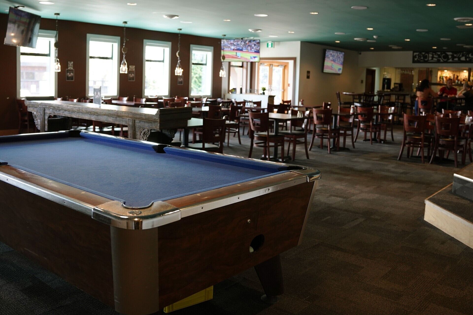A pool table in a room with multiple tables and chairs, large windows, and a bar area.