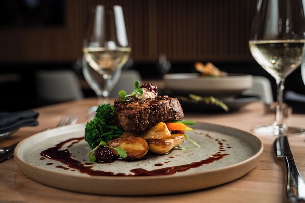 Un plato de comida con un bistec, verduras y salsa, sobre una mesa con una copa de vino y cubiertos.