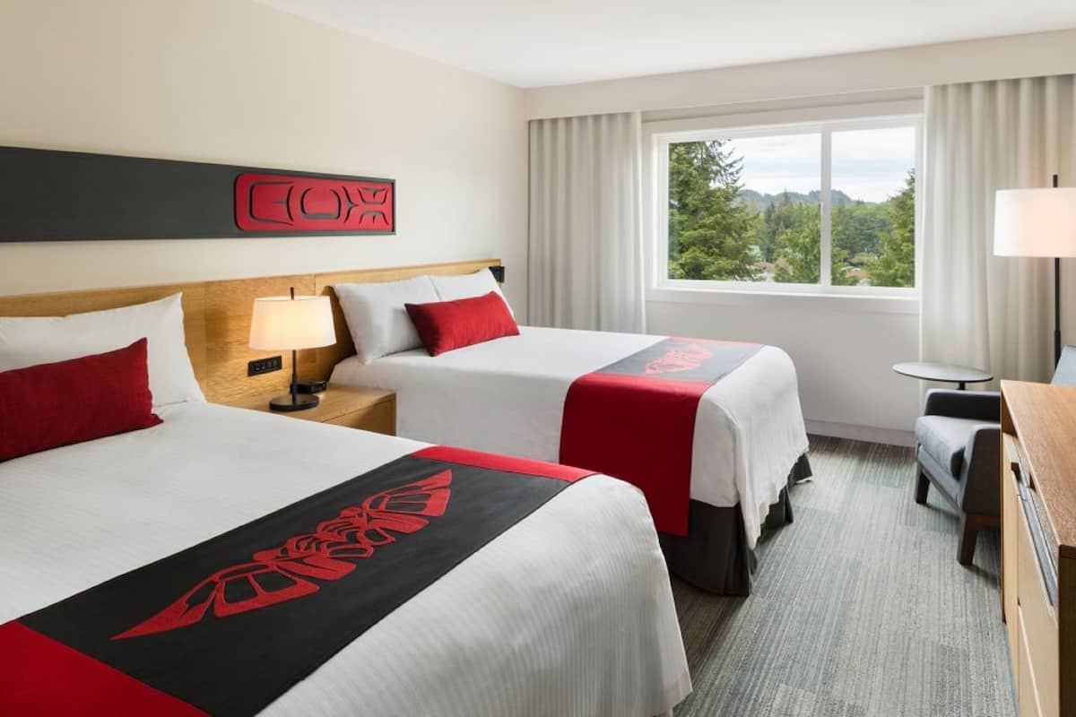 A hotel room with two beds, a nightstand, a desk, and a window with a view of greenery.