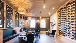 Un interior moderno con una pared larga de botellas de vino, una zona de estar con un banco y sillas, e iluminación colgante.